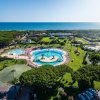Vista dall'alto di un complesso con piscine circolari, prato, e mare sullo sfondo.