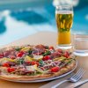 Pizza con verdure e prosciutto su un tavolo, birra e acqua, vista piscina.