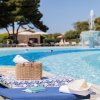 Lettino da piscina con borsa, cappello, e libro, a bordo vasca. Sullo sfondo, fontana e ombrelloni.