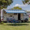 Camper parcheggiato su prato, sotto una tenda blu a strisce, con persone sedute su sedie a sdraio.