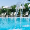 Dettaglio di getti d'acqua in una piscina, con lettini e ombrelloni bianchi sullo sfondo.